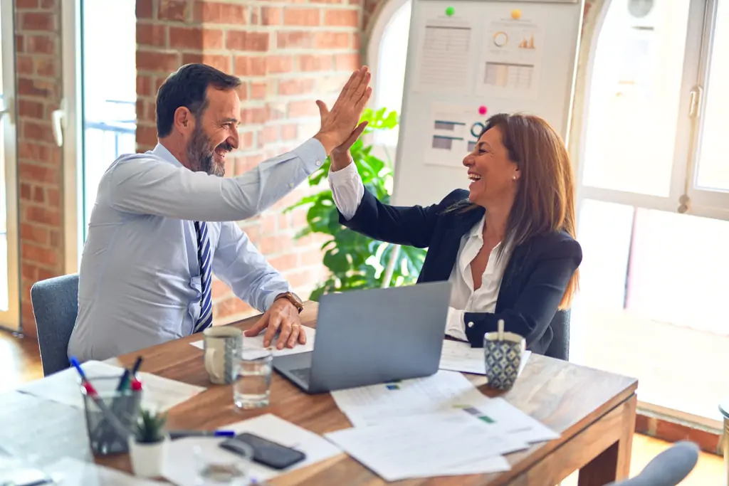 A businessman and businesswoman sitting at a table with work documents, giving each other a highfive.