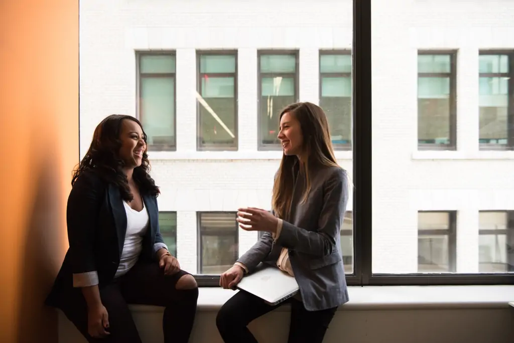 Two businesswomen sitting on a windowsill, laughing.